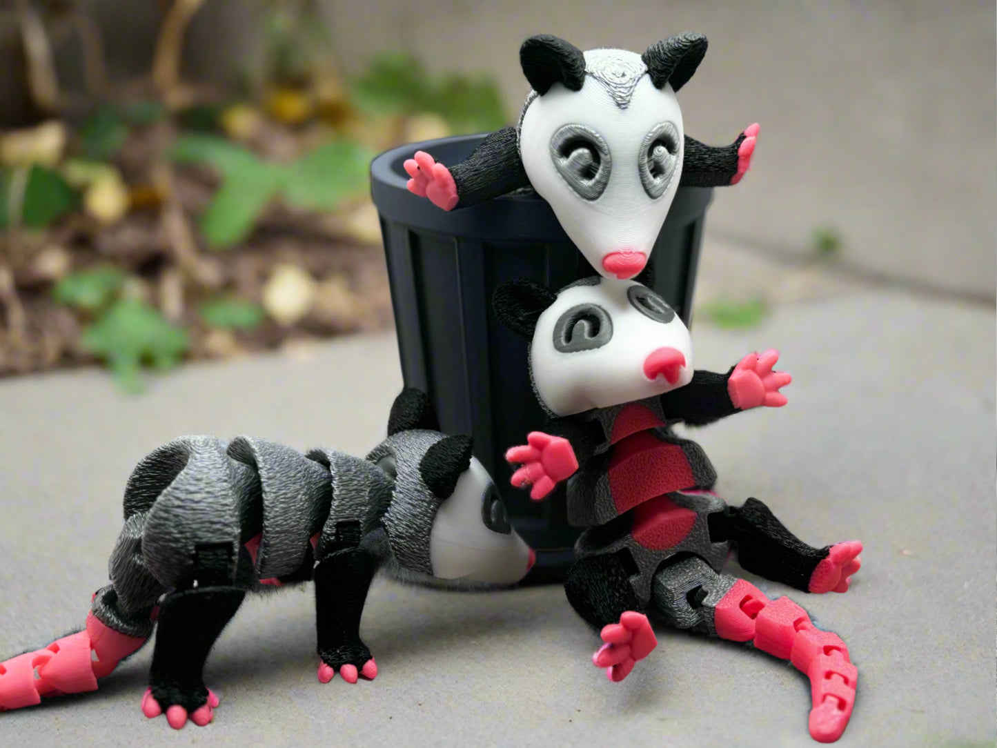 Three 3D-printed opossums are shown in playful poses. One is perched on a mini trash can, another lies on its back, and the third appears to crawl forward. They have white faces, black eyes, and pink noses, ears, and feet, with gray and black bodies.